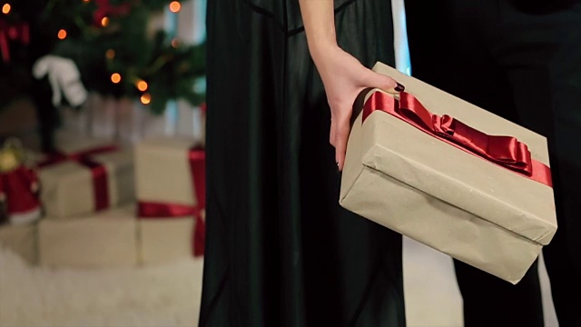 身着黑色晚礼服的女人手里拿着一件带红丝带的礼物，一个穿着黑色西装的男人在一个有壁炉和圣诞树的白色房间里视频下载