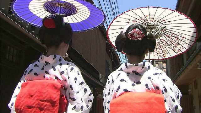 两个舞妓穿着浴巾，撑着雨伞走在一条狭窄的街道上。视频素材