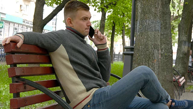 一个年轻人坐在城市公园的长椅上，正在讲电话视频素材