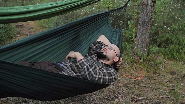 夏日的绿色森林里，人们在吊床上休息。留胡子戴眼镜的男人视频下载