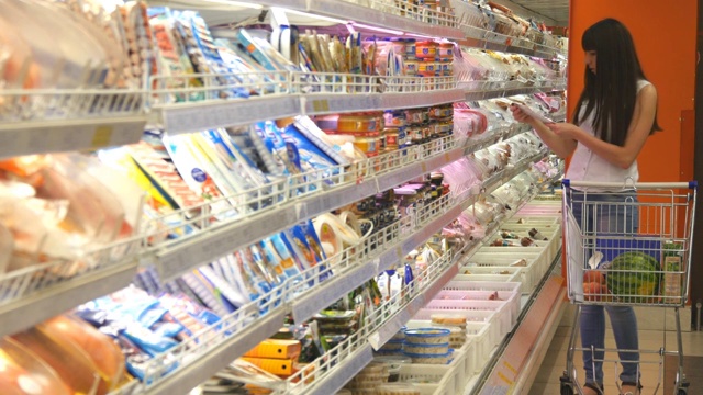 年轻女子推着购物车在超市的冷藏区购买乳制品或冷藏食品。一个女孩走到商店的冰箱前，从里面拿东西视频下载