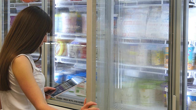 年轻女子在超市的冷藏区打开玻璃门购买乳制品或冷藏食品。女孩在商店从冰箱里拿东西，用平板电脑检查购物清单视频素材