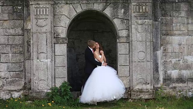 可爱的新郎站在古老的拱门上，拥抱着美丽的新娘，温柔地亲吻她视频下载