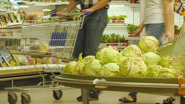 一对年轻夫妇推着超市手推车走在购物中心的杂货店里。女人坐下来，挑白菜放进篮子里视频素材
