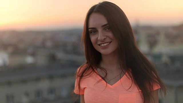 性感的长发女孩在镜头前转过身微笑，背景是夕阳下模糊的城市视频下载