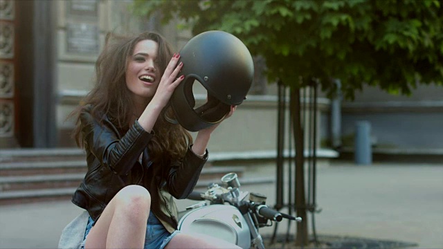 一个美丽的白人女孩骑摩托车的城市肖像。穿着皮夹克的黑发美女，脱下摩托车头盔，然后笑了视频下载