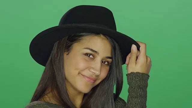 年轻女子戴着一顶帽子，调皮地摆姿势，在一个绿色屏幕的工作室背景视频下载