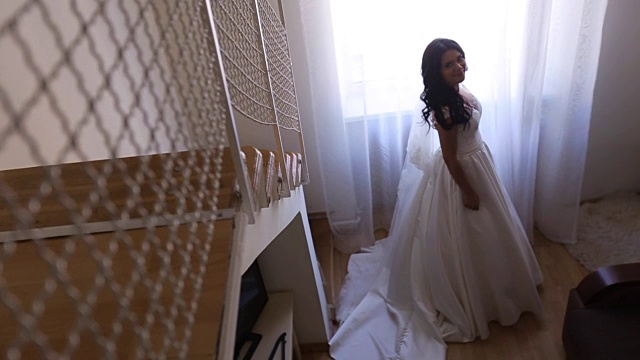 时尚豪华华丽的黑发新娘摆pose靠近窗户的背景酒店房间。新娘花束。前视图。视频下载