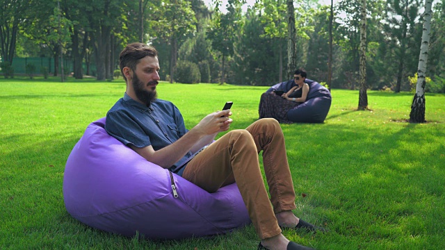一个穿着蓝色衬衫的年轻人坐在公园的椅子上，通过电话获得了一条信息视频下载