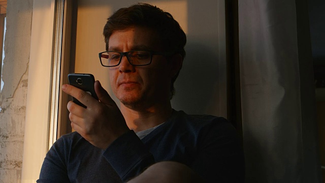 悲伤的男人看着日落的窗户，手里拿着手机视频素材