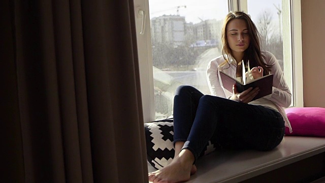 迷人的时尚的年轻女孩在窗台上看书和透过窗户看视频下载