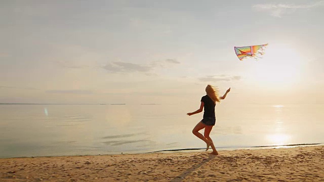 无忧无虑的年轻女子在海滩上跑着放风筝。映衬着美丽的晚霞天空视频素材