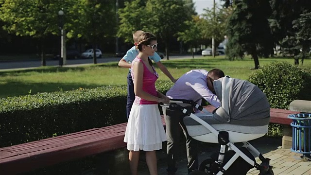 年轻的妈妈和爸爸正推着婴儿车站在公园的长椅旁。视频下载