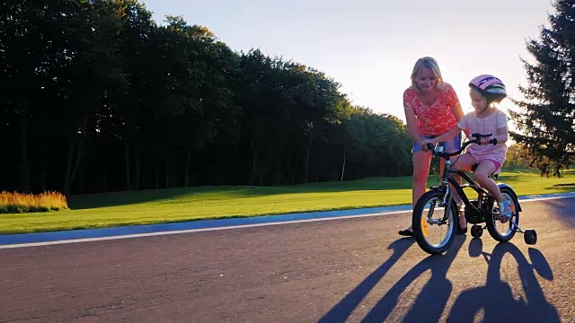 妈妈和女儿在公园里玩。他教她在夕阳的背景下骑自行车视频素材