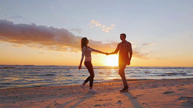 斯坦尼康慢动作镜头:浪漫的夫妇漫步在日落的海滩上，手牵着手，度过美好的时光视频素材