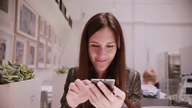 一个穿着皮夹克，黑头发的漂亮年轻女人用着她的智能手机，说话，微笑。墙上的画视频素材