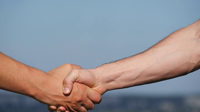 在蓝天背景下，两个肌肉发达的白人男子友好地握手。在室外摇晃男性手臂。两个强壮的男人在外面紧紧握手。团队合作和友谊。特写镜头视频下载