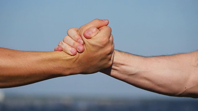 在蓝天背景下，两个肌肉发达的白人男子友好地握手。在室外摇晃男性手臂。两个强壮的男人在外面紧紧握手。团队合作和友谊。特写镜头视频下载