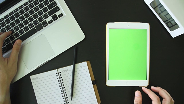 一个人在笔记本电脑和白色平板电脑上工作的俯视图，桌面上有一个空白的绿色触摸屏。视频素材