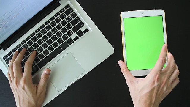 一个人在笔记本电脑和白色平板电脑上工作的俯视图，桌面上有一个空白的绿色触摸屏。视频素材