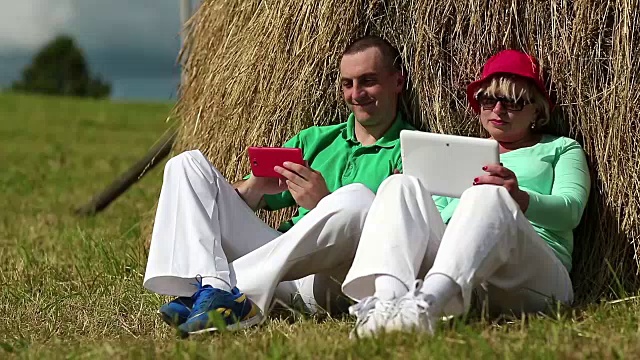 一个拿着红色智能手机的男人和一个拿着平板电脑的女人坐在干草堆旁边视频下载