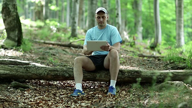 一名男子带着平板电脑坐在森林里的一根原木上视频素材