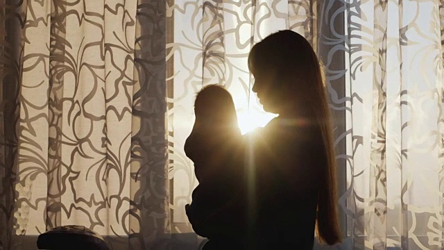 剪影母亲与婴儿。日落时，站在窗前，阳光明媚地照耀着视频下载