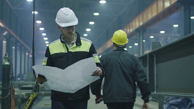 戴安全帽的工程师正在带着蓝图穿过一个重工业工厂。视频素材
