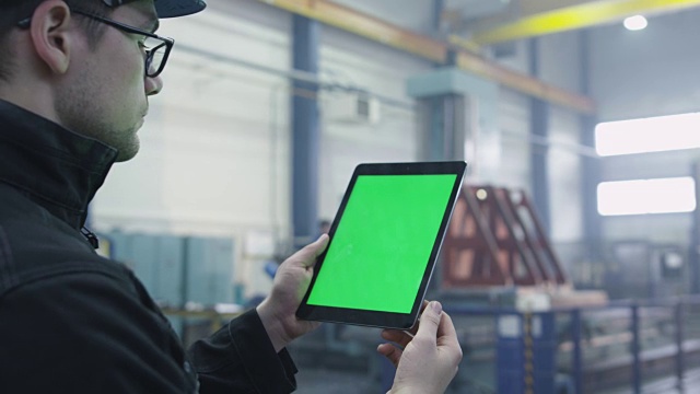 在一个工厂的工业环境中，一个工人正在使用一个带有绿色模拟屏幕的平板电脑的镜头。视频素材
