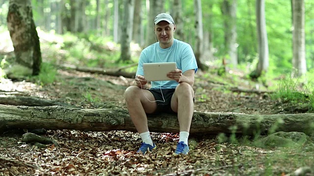 一名手持平板电脑的男子坐在森林里倒下的一棵树上视频素材