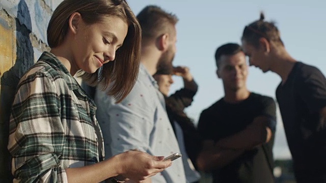 迷人的少女使用手机旁边的一群她的青少年朋友在户外城市环境。视频素材