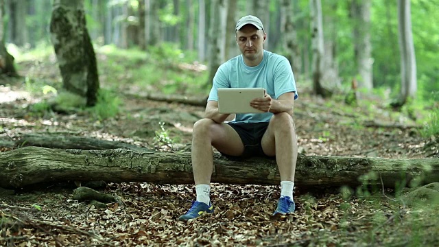 一名手持平板电脑的男子坐在森林中的一根木头上视频素材