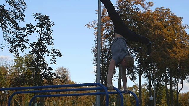 强壮的肌肉男在公园里做倒立。健美的肌肉男在户外的单杠上表演特技。运动员训练时在肌肉外做一个倒立。慢动作视频下载