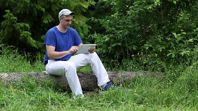 一名手持平板电脑的男子坐在森林里倒下的一棵树上视频素材
