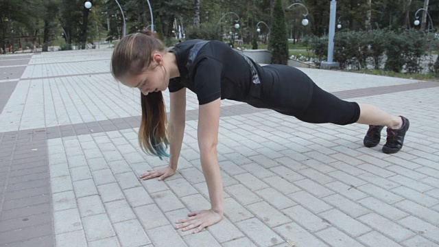 一个穿着运动服的小女孩在公园里做俯卧撑，双手放在地上。在铺着石板的地面上视频下载