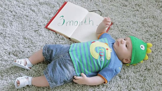 3个月大的婴儿戴着绿龙形状的帽子躺在灰色的地毯上。视频下载