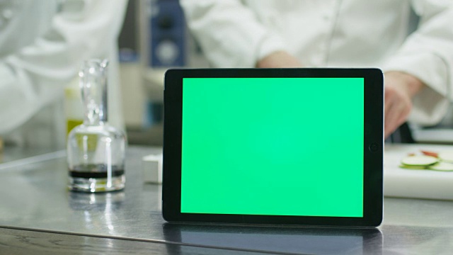 在一间商业厨房的桌子上放着一台绿色屏幕的平板电脑，背景是厨师正在准备食物。视频素材