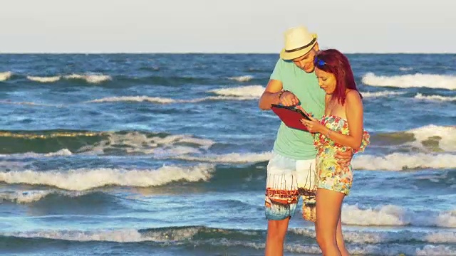 年轻夫妇有一个夏天的一天在海边与平板电脑的乐趣视频素材