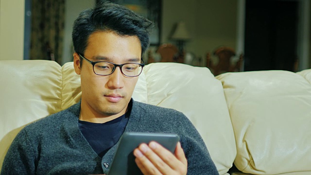 一个戴眼镜的亚洲年轻人。他坐在家里的沙发上看电子书视频素材