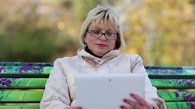 一个金发女人坐在公园的长椅上，用着白色的平板电脑视频素材