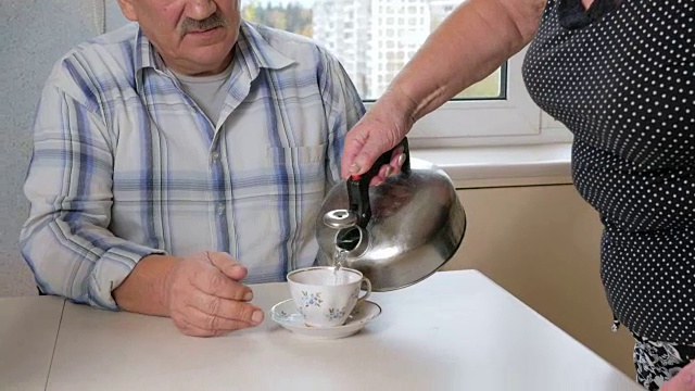 一个上了年纪的女人在家里的厨房里给丈夫沏茶。她把杯子放在桌子上，倒了热水。护理的概念视频下载
