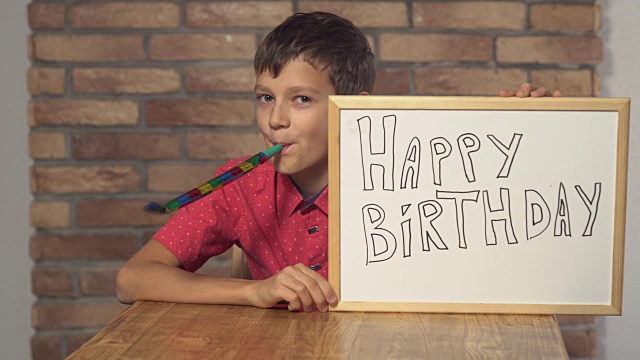 孩子坐在书桌前，手里拿着写着生日快乐的挂图，背景是红色的砖墙视频素材