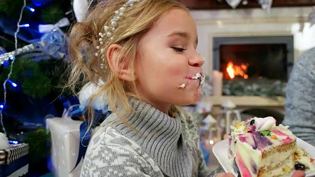 女孩愉快地吃一大块蛋糕在圣诞树，家人在壁炉边吃甜点与茶，妈妈和女儿享受在一起的时间视频素材