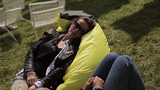两个戴着太阳镜的女孩在绿色草地上的黄色沙袋上放松，微笑着。夏天的节日。休息视频下载