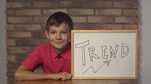 孩子坐在桌前，拿着写有字母趋势的挂图，背景是红砖墙上视频素材