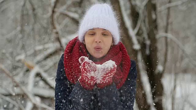 年轻的姑娘戴着白色的针织帽，戴着红色的围巾和手套吹雪视频素材