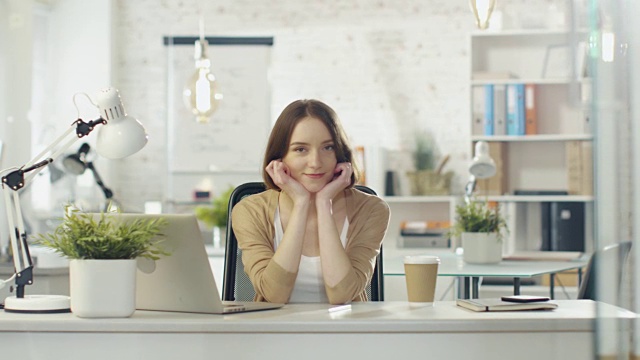 肖像拍摄的一个美丽的年轻女孩坐在她的办公桌在一个明亮的办公室。她对着镜头迷人地微笑。视频素材