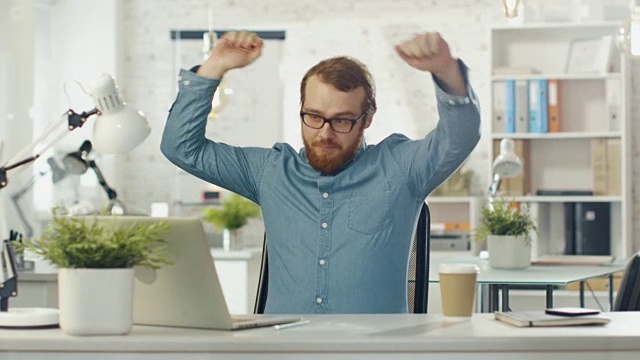 在明亮的办公室里，蓄着胡子的科技男坐在他的办公桌前跳舞。视频素材