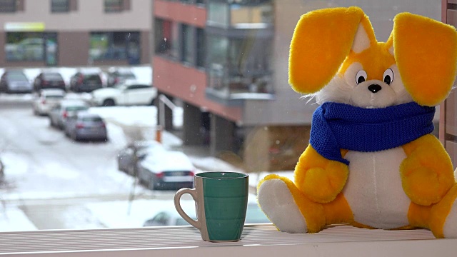 冬天，黄色的玩具兔子拿着茶杯坐在窗边。雪花下降。视频下载