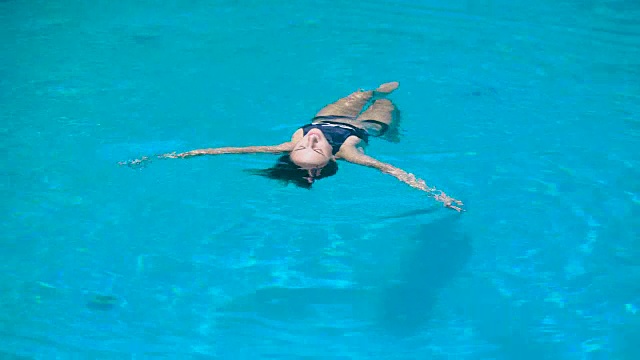可爱的fit女人在黑色泳衣是放松luying在水上。年轻的身材匀称的女孩在游泳池里飘浮着舒适和快乐视频下载